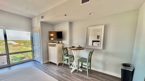 A modern hotel room with a large window, a flat-screen TV, a small dining table, and a trash bin.