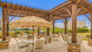An outdoor patio area with tables and chairs, a pergola with a thatched roof, and stone pillars.