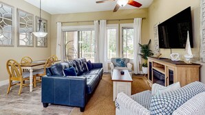 A spacious living room with a blue sofa, a white patterned sofa, a wooden dining table with chairs, a ceiling fan, and a TV mounted on the wall. - Ground-floor condo with patio, central AC, shared pool, & tennis (Miramar Beach)