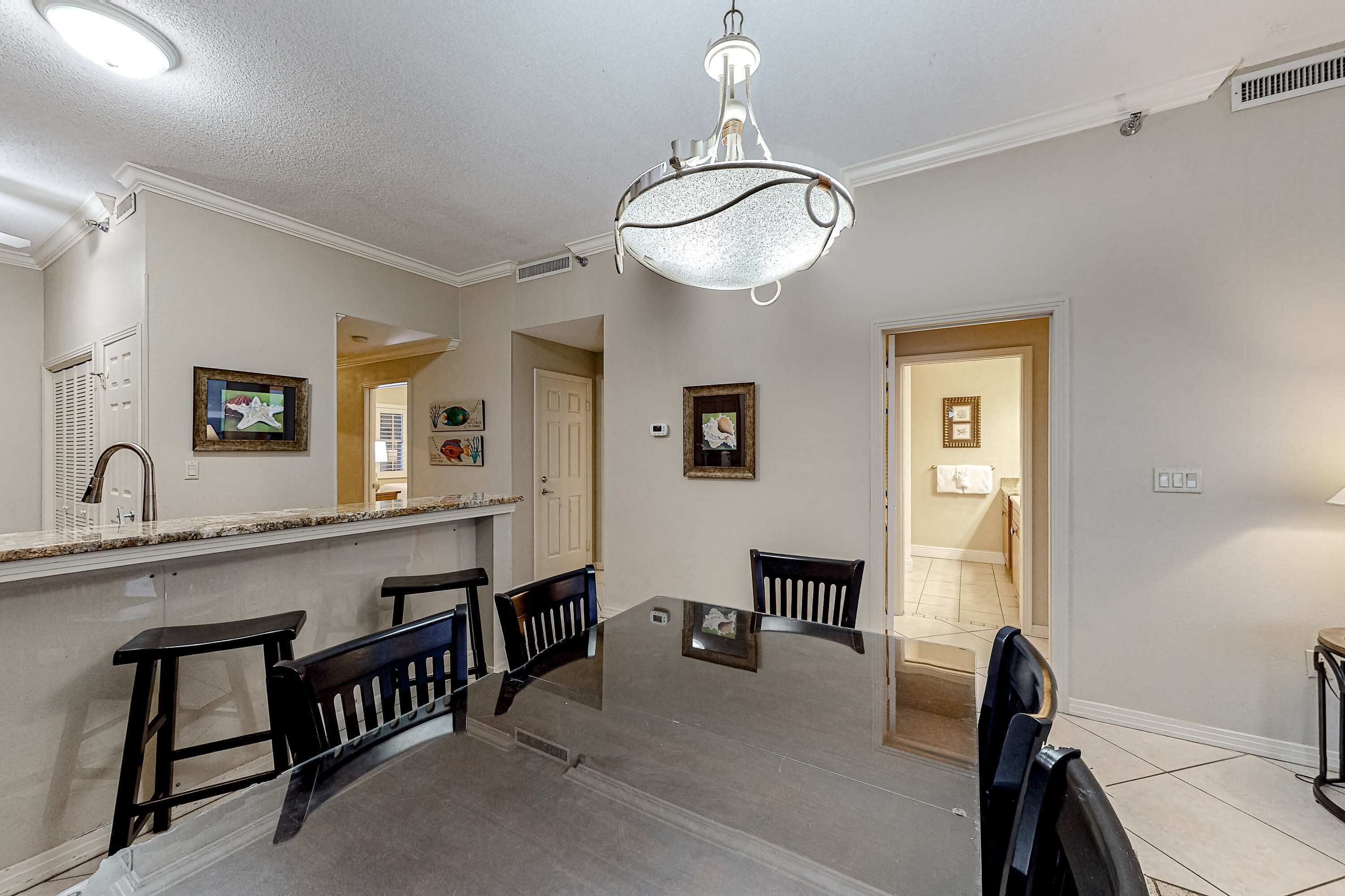 A dining area with a glass table and four chairs, a chandelier, and a kitchen counter with a sink and cabinets.