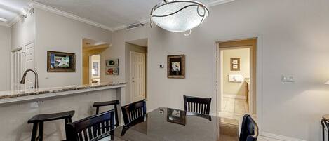 A dining area with a glass table and four chairs, a chandelier, and a kitchen counter with a sink and cabinets.