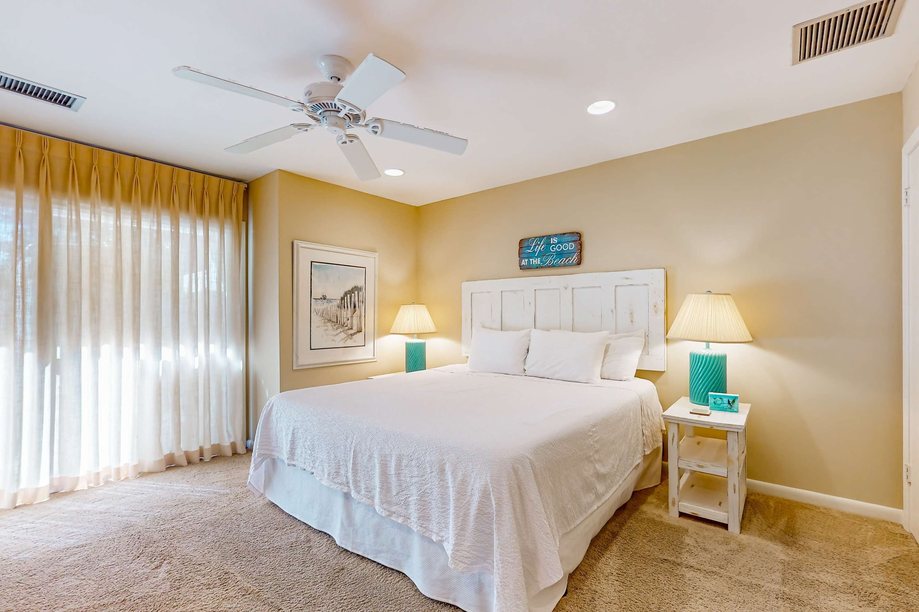 A hotel room with a large bed, two bedside lamps, a ceiling fan, and a beach-themed sign on the wall.