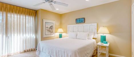 A hotel room with a large bed, two bedside lamps, a ceiling fan, and a beach-themed sign on the wall.