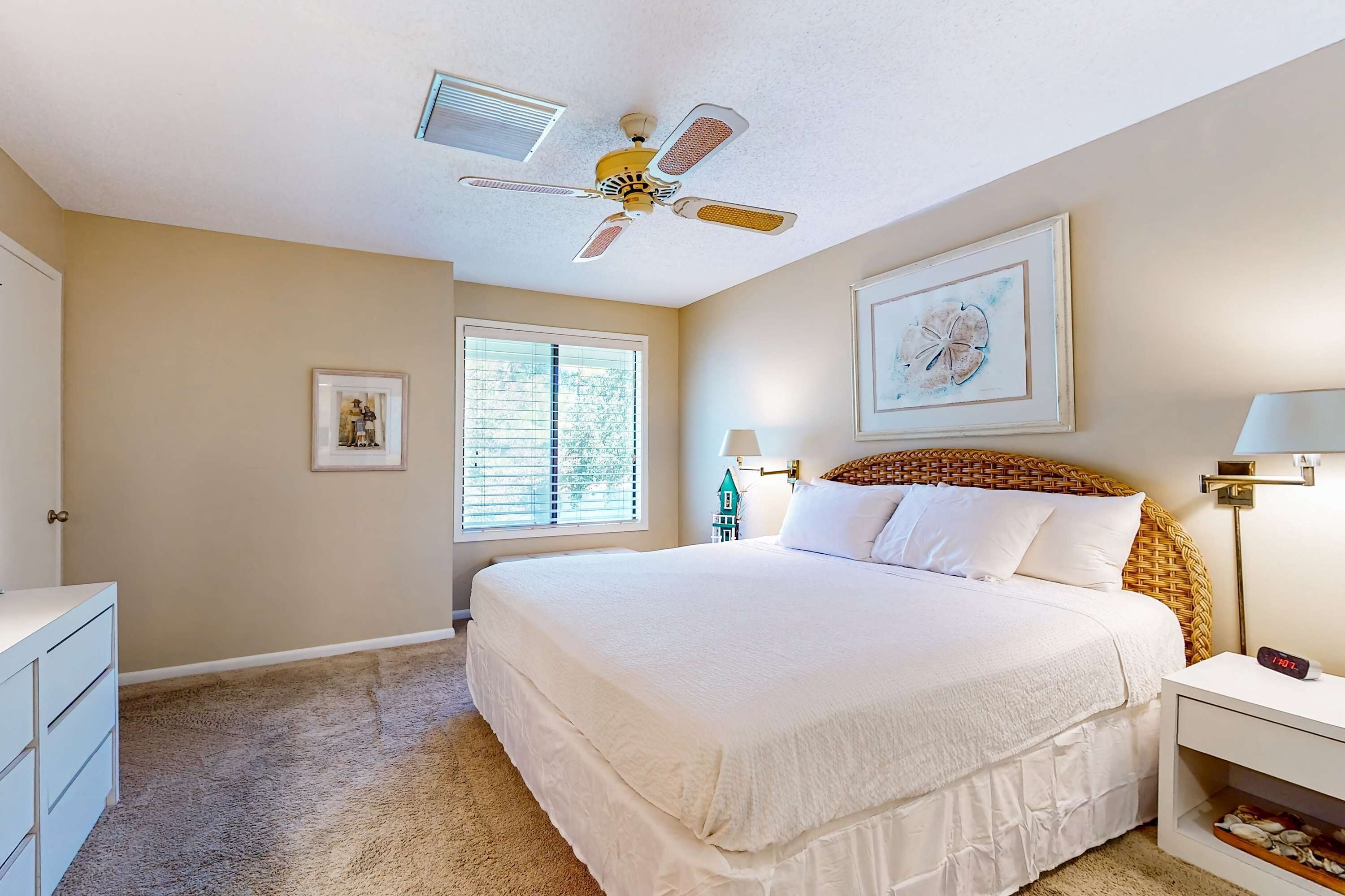 A bedroom with a large bed, a ceiling fan, a window with blinds, a dresser, and a framed picture on the wall.