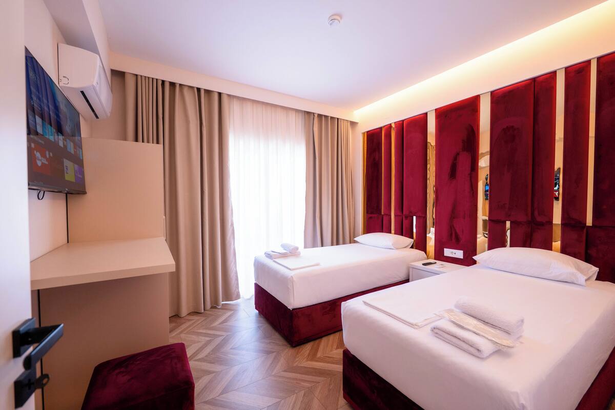 A hotel room with two beds, a desk, a chair, and a large window with curtains.