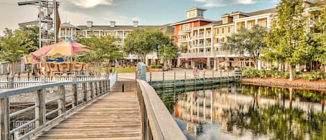 A wooden boardwalk over a calm water body, leading to a row of multi-story buildings with balconies, a carousel, and a clear sky.