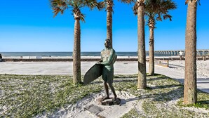 Una statua raffigura una persona che tiene una tavola da surf, circondata da palme, con una spiaggia e l'oceano sullo sfondo.