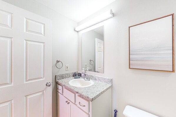 Une salle de bain avec un lavabo blanc, des armoires roses, un grand miroir et une œuvre d’art encadrée accrochée au mur.