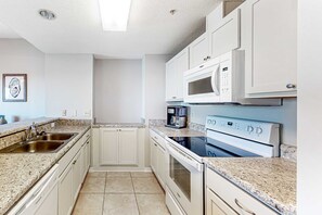 A compact kitchen with white cabinets, a double sink, a microwave, and a stove. - Sunlit & tropical beachfront condo with pool, gym, balcony views (Pensacola Beach)