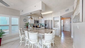 Cocina moderna con una isla central, sillas blancas, electrodomésticos de acero inoxidable y un amplio ventanal con persianas.