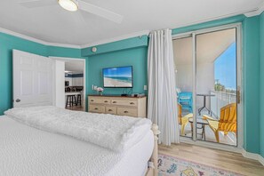 A bedroom with a bed, a dresser, a TV, and a balcony with chairs. - Gulf-front home with balcony, ocean views, pool and beach access (Fort Walton Beach)