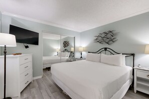 A bedroom with a large bed, a bedside table with a lamp, a dresser, a flat-screen TV mounted on the wall, and a mirror. - Sunlit Gulf-front condo with gorgeous water views, pool, balcony, & tennis court (Fort Walton Beach)