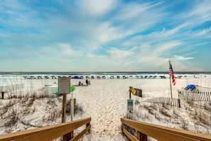 A beach with a wooden boardwalk leading to the shore, blue umbrellas, and an American flag. - Fifth floor oceanfront condo with pool, ocean view, W/D (Fort Walton Beach)