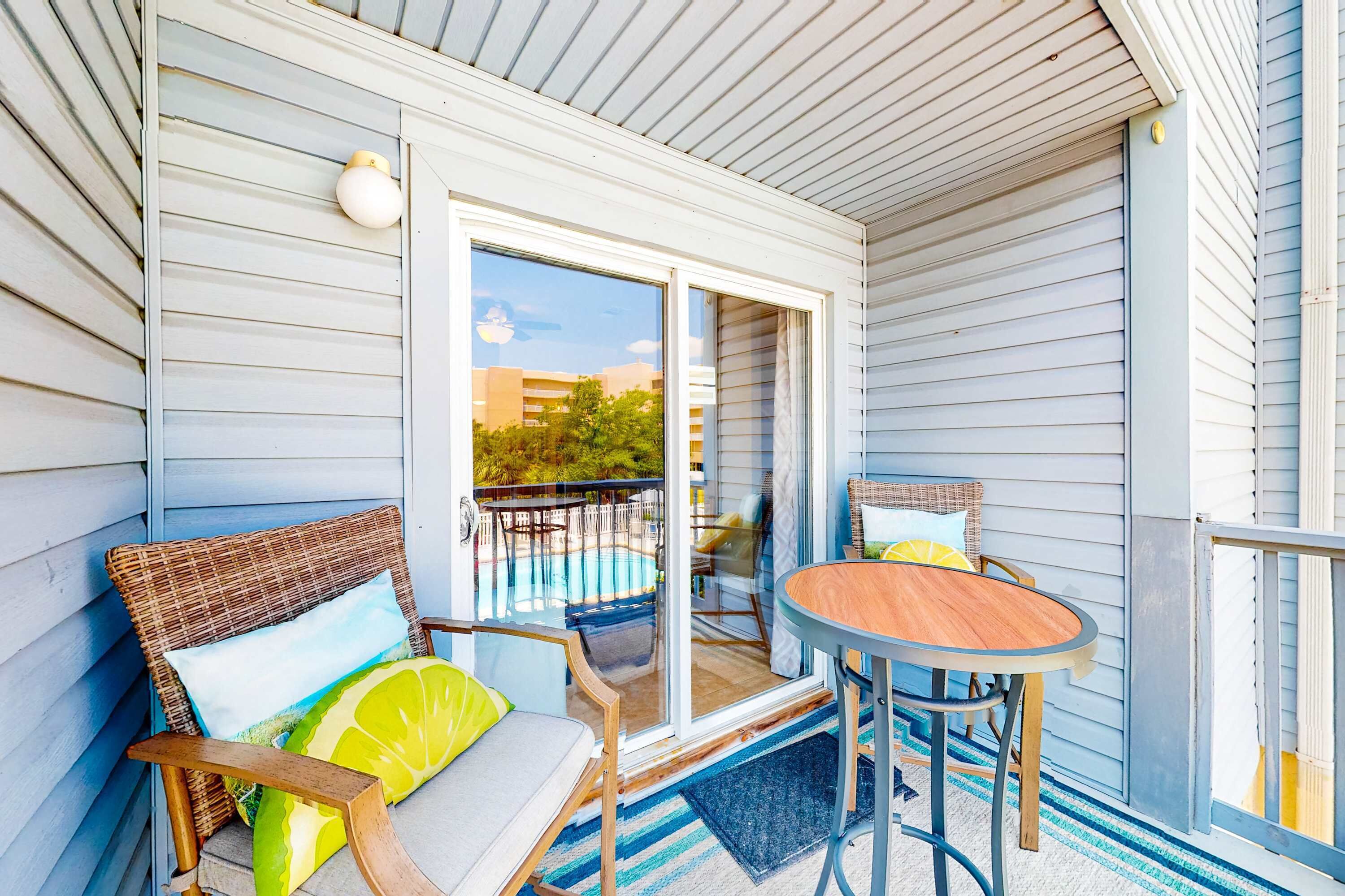 A covered porch with wicker furniture, a wooden table, and a view of a swimming pool and buildings in the background.
