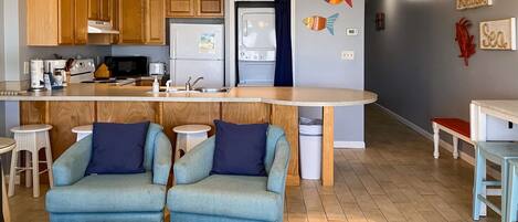 Une cuisine moderne avec des meubles en bois, un îlot central intégré et un coin salon avec deux fauteuils.