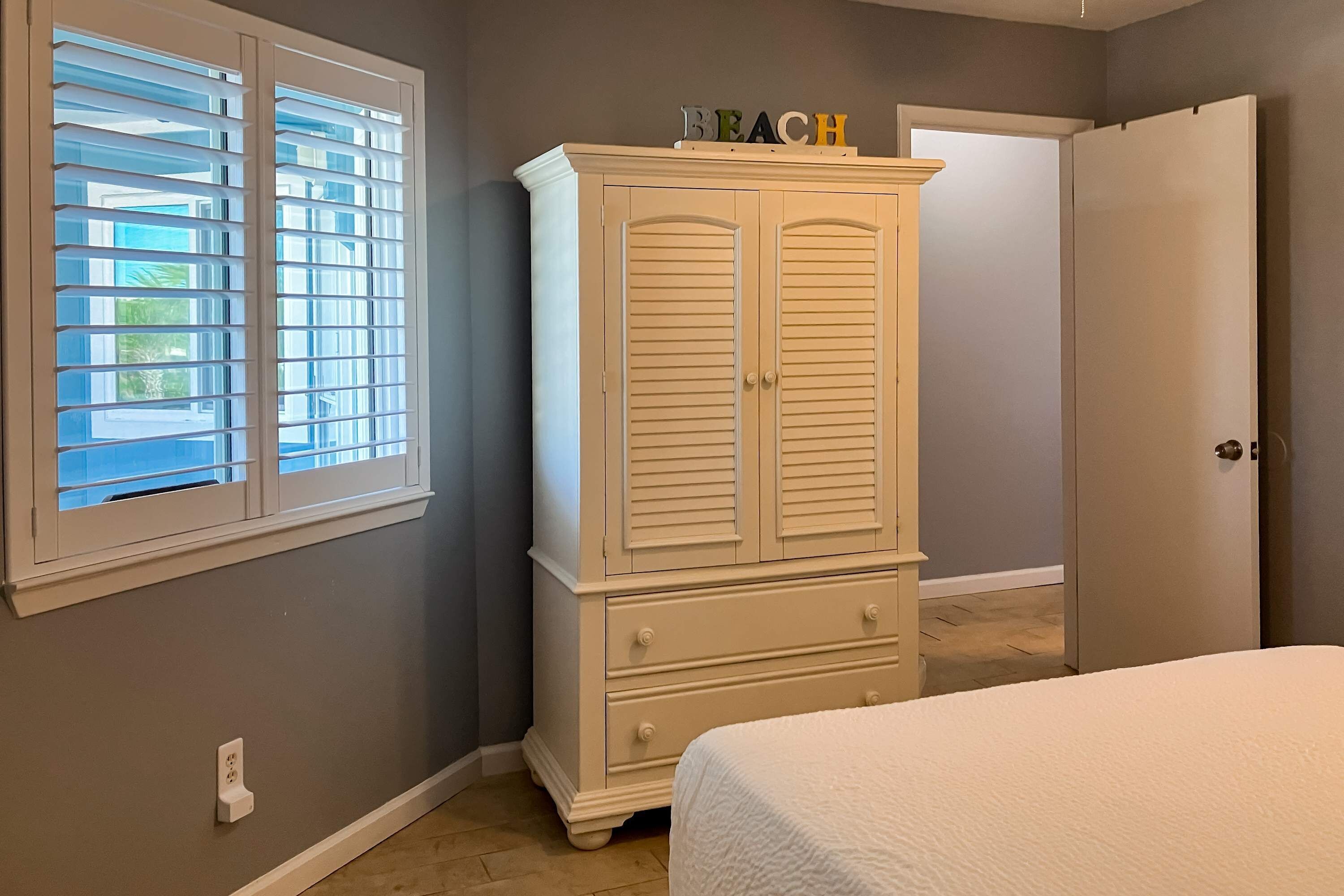 A bedroom with a white bed, a white dresser with drawers, a window with blinds, and a door.