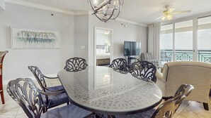 Een eetgedeelte met een ronde glazen tafel, zwarte metalen stoelen, een kroonluchter en een televisie op de achtergrond.