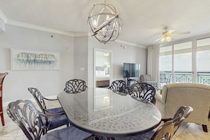 A dining area with a round glass table, black metal chairs, a chandelier, and a TV in the background. - Gulf Front Condo Pools Onsite, Nearby Activities, Shops, Restaurants (Fort Walton Beach)
