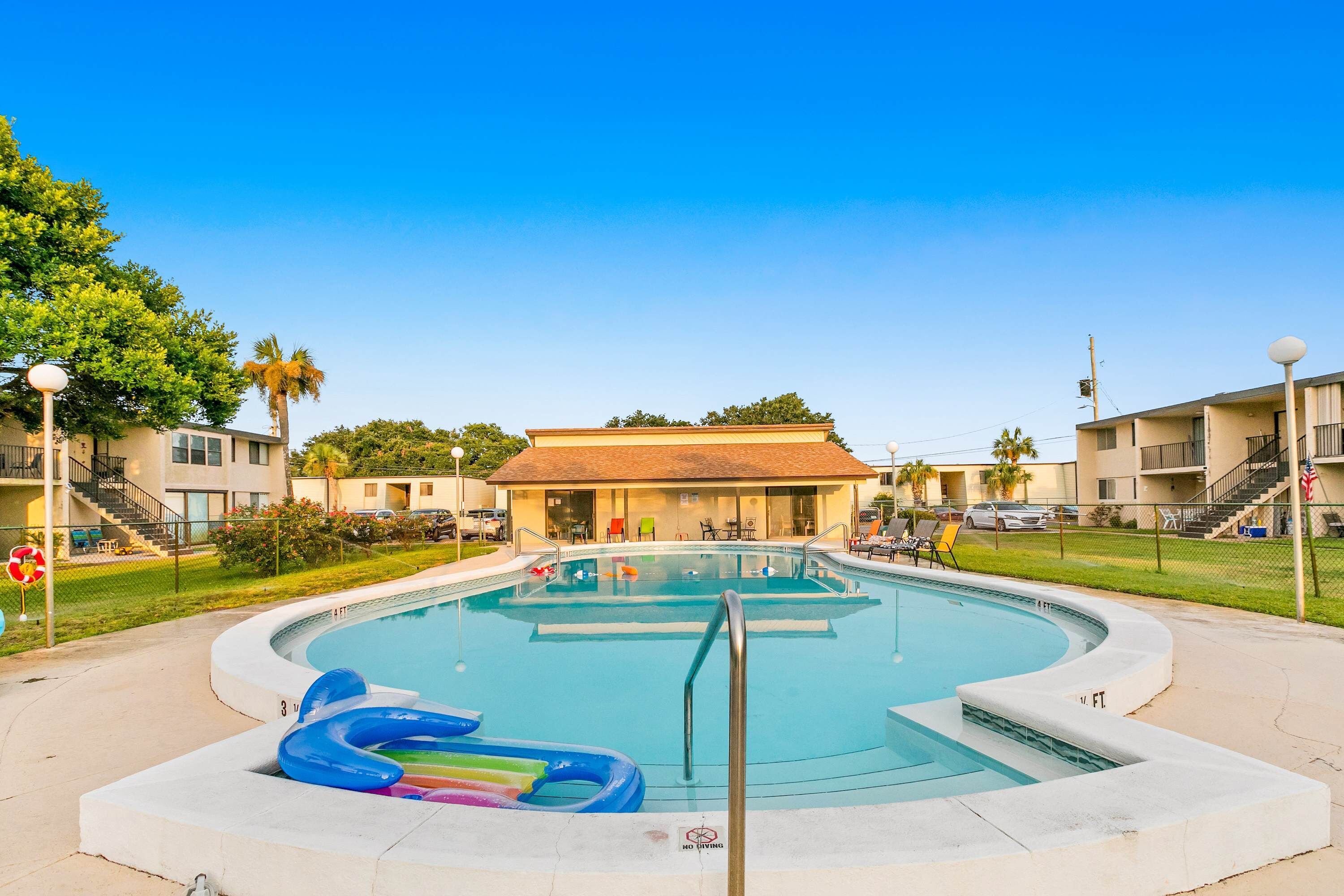 Una piscina con una escalera metálica, rodeada por una solana de hormigón. Hay sillones de descanso y una palmera al fondo.