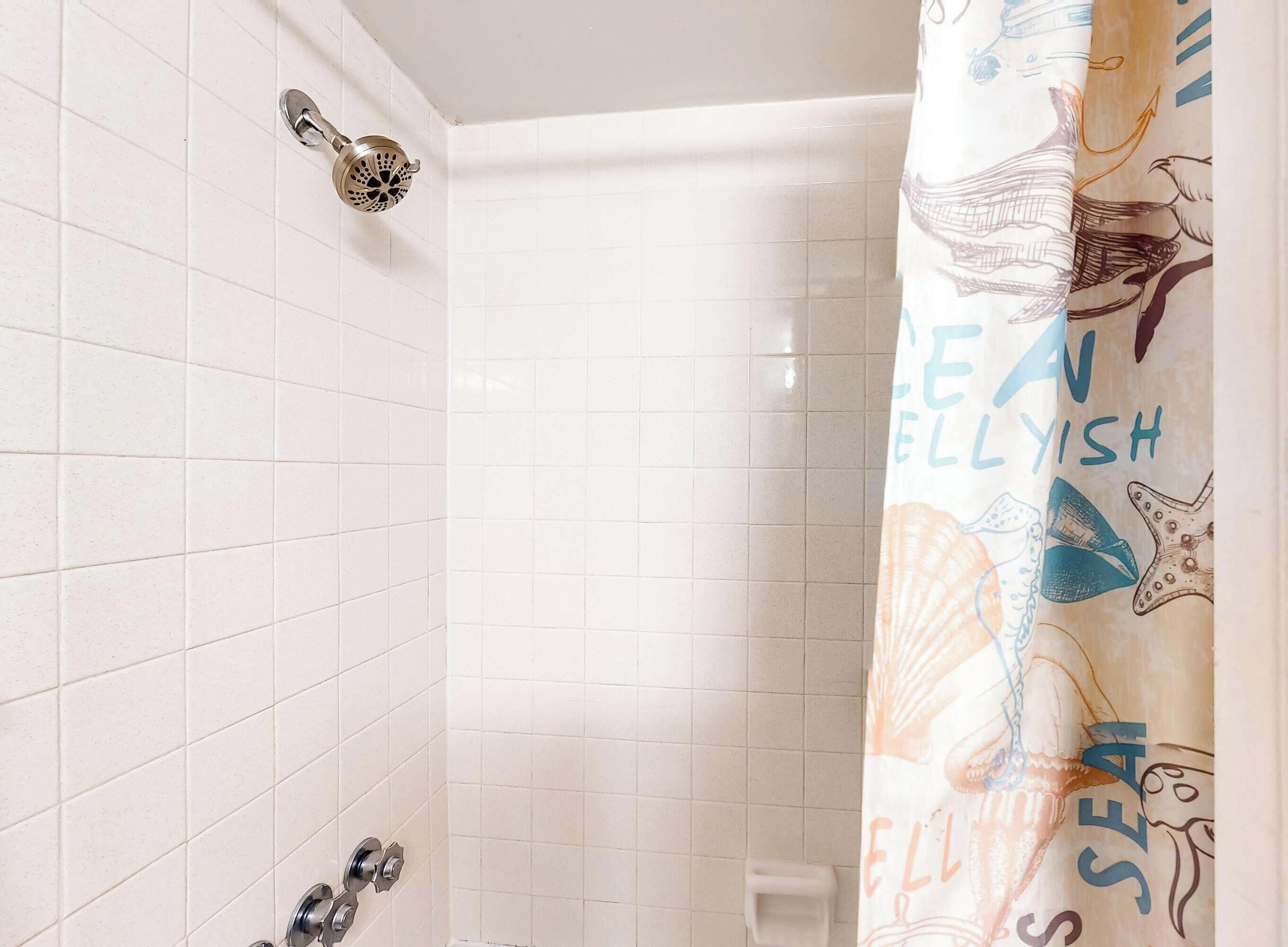 Une salle de bain avec des murs carrelés de carreaux blancs, une douche avec un pommeau de douche amovible et un rideau de douche orné de motifs marins.