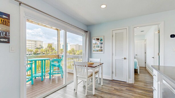 Een balkon met turquoise stoelen en een houten tafel, een witte deur met een prikbord, en een uitzicht op gebouwen door de glazen deuren.