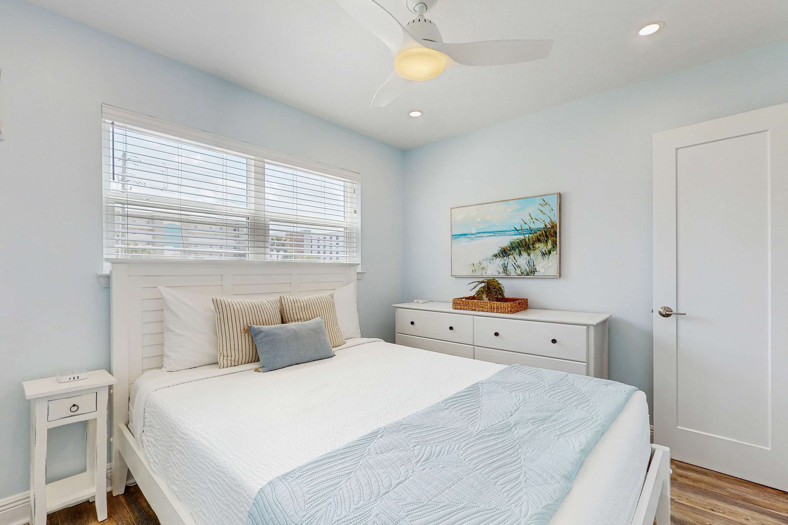 A bedroom with a bed, a nightstand, a dresser, a window with blinds, a ceiling fan, and a painting on the wall.