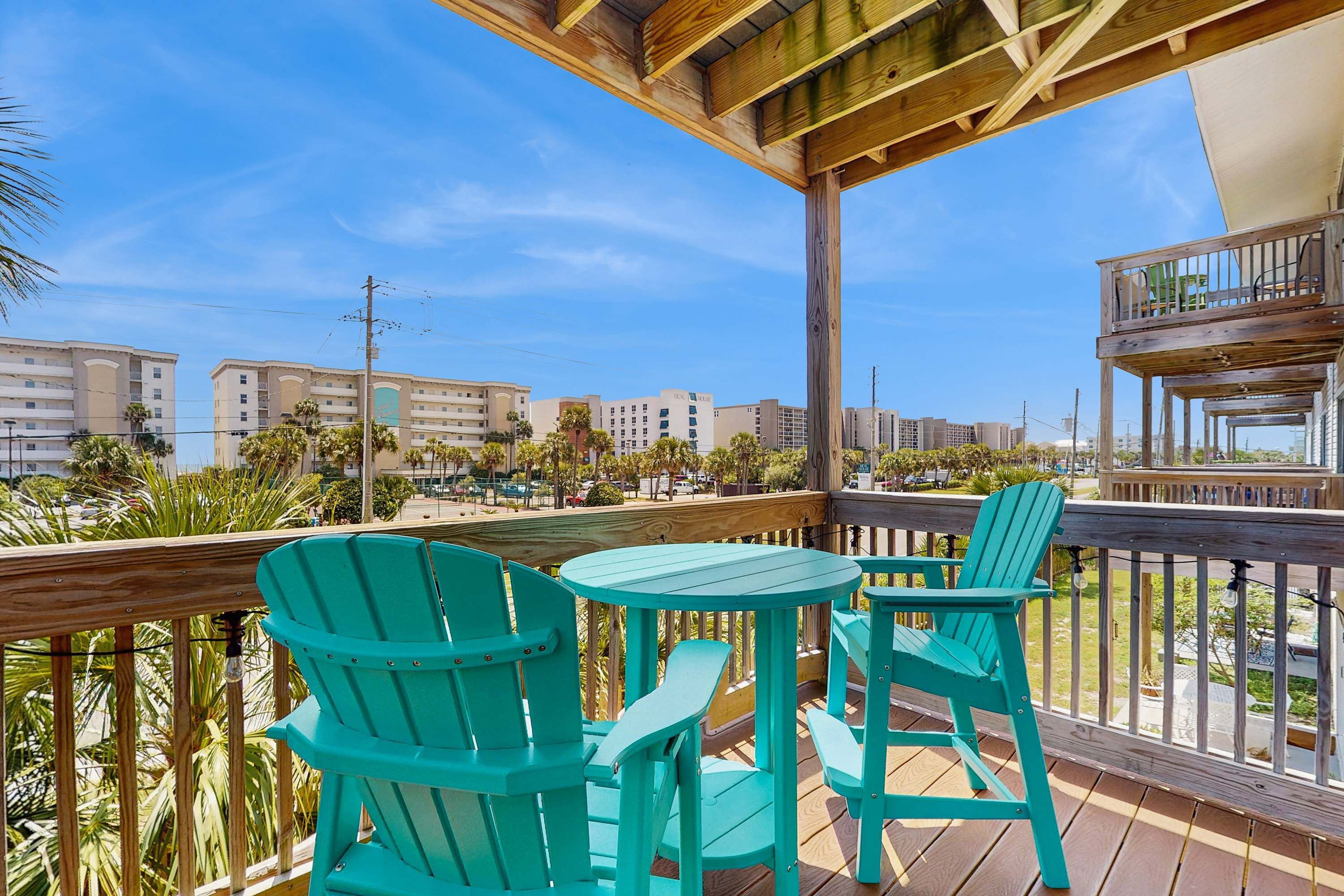 A wooden deck with turquoise chairs and a table, overlooking a beachfront with buildings and palm trees.