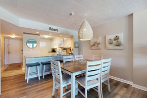 Un coin repas avec une table en bois et des chaises blanches, un luminaire suspendu et une cuisine en arrière-plan.