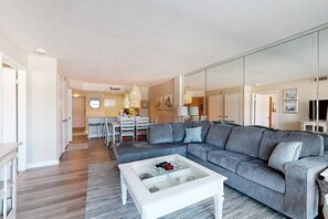 Un salon moderne avec un canapé d’angle gris, une table basse en verre et un coin repas à l’arrière-plan.