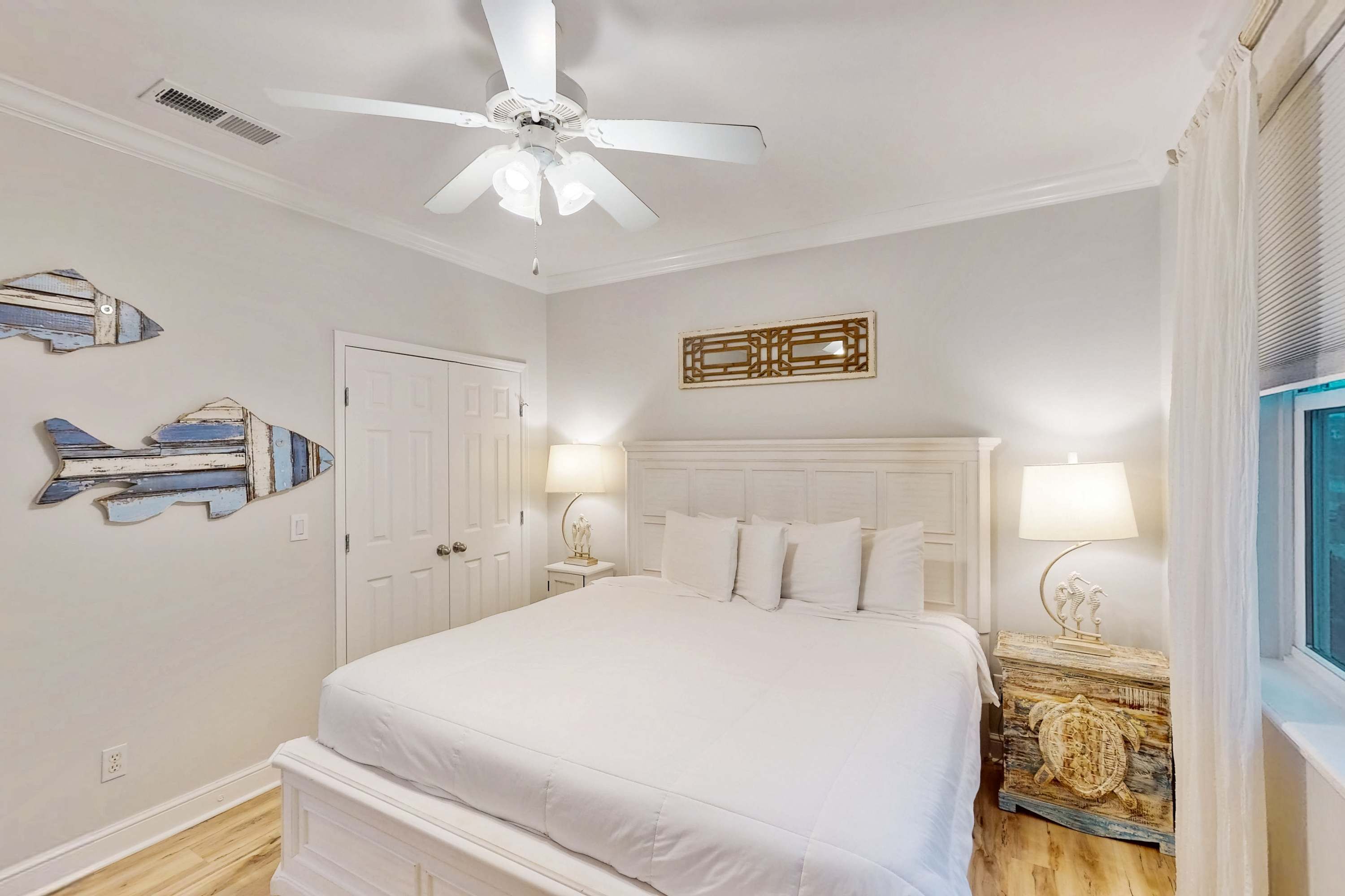 A bedroom with a white bed, a ceiling fan, a wooden nightstand with a lamp, and decorative wall art.