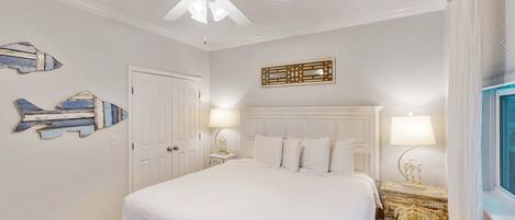 A bedroom with a white bed, a ceiling fan, a wooden nightstand with a lamp, and decorative wall art.