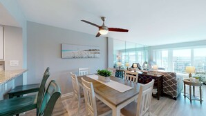 A modern living room with a dining area, a ceiling fan, a large window, and a city view. - Sea Oats 708 - 7th floor condo in beachfront complex with pool, Gulf view, W&D, & fast WiFi (Fort Walton Beach)