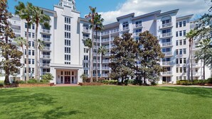 Um hotel com vários andares e entrada central, rodeado por um gramado bem cuidado e palmeiras.