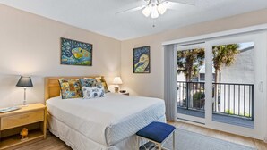 Ein Schlafzimmer mit einem großen Bett, einem Nachttisch, einer Bank, zwei Gemälden und einem Balkon mit Blick auf Palmen.