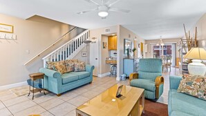 Ein großzügiges Wohnzimmer mit einem blauen Sofa, einem Glastisch und einem blauen Sessel. Eine Treppe führt zu einer oberen Ebene.