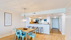 En rummelig spiseplads med et træbord og blå stole, et køkken i baggrunden og et indramet citat på væggen.