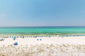 A beach with white sand and clear turquoise water. There are numerous blue umbrellas and beach chairs arranged along the shoreline. - Seventh floor Gulf-front suite with amazing views, washer&dryer, AC & pool (Fort Walton Beach)