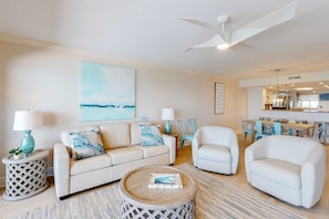 A living room with a beige sofa, two armchairs, a coffee table, and a ceiling fan. There is a painting on the wall and a kitchen area in the background. - Seventh floor Gulf-front suite with amazing views, washer&dryer, AC & pool (Fort Walton Beach)