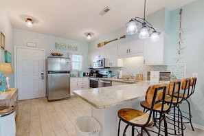 A kitchen with white cabinets, a stainless steel refrigerator, a marble island with bar stools, and a 'BEACH HOUSE' sign. - Condo with partial bay view, onsite fishing dock, beach access, & shared pool (Pensacola Beach)