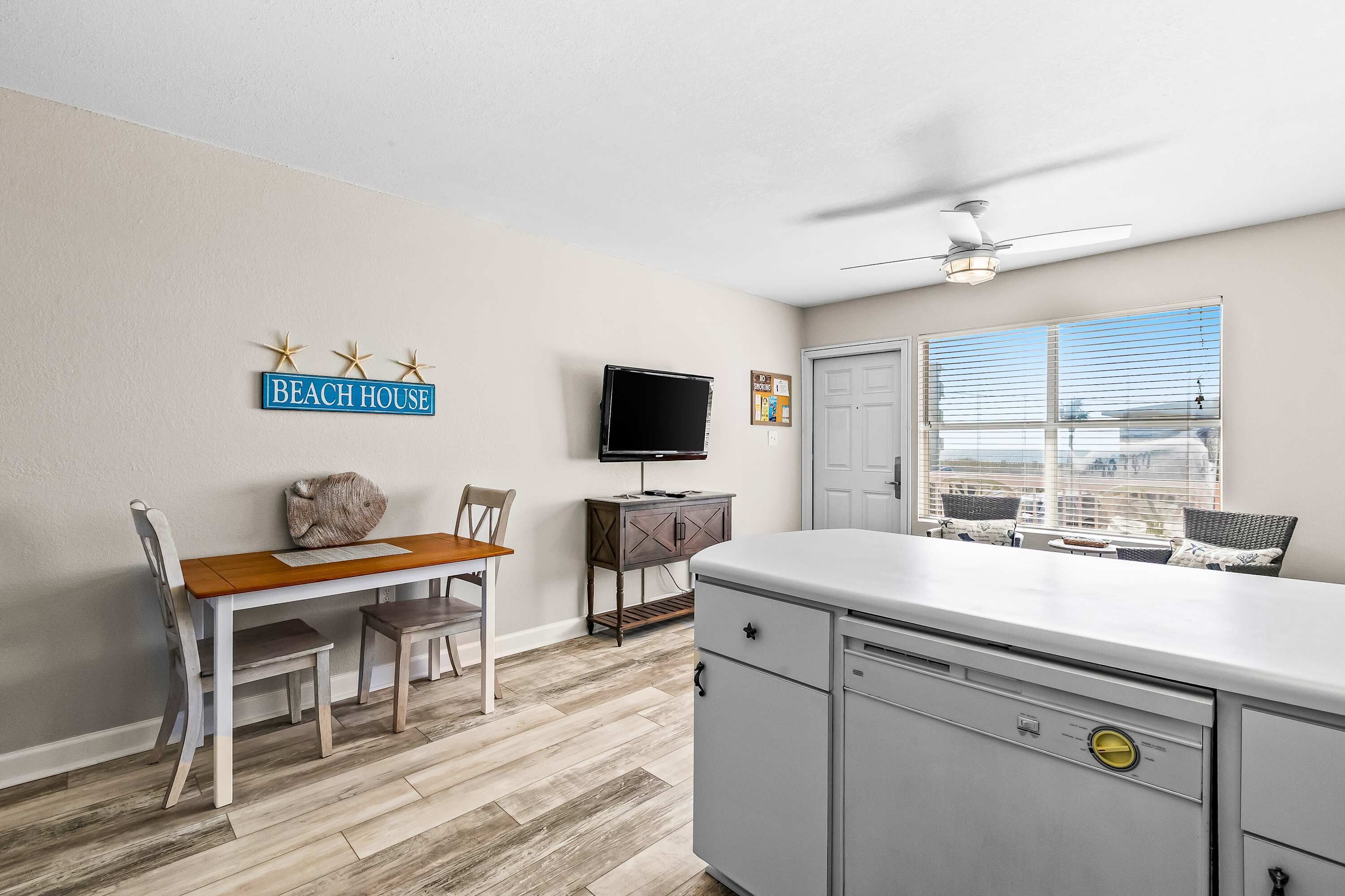 A kitchen with a white countertop, grey cabinets, and a ceiling fan. There is a dining table with chairs and a television on the wall.