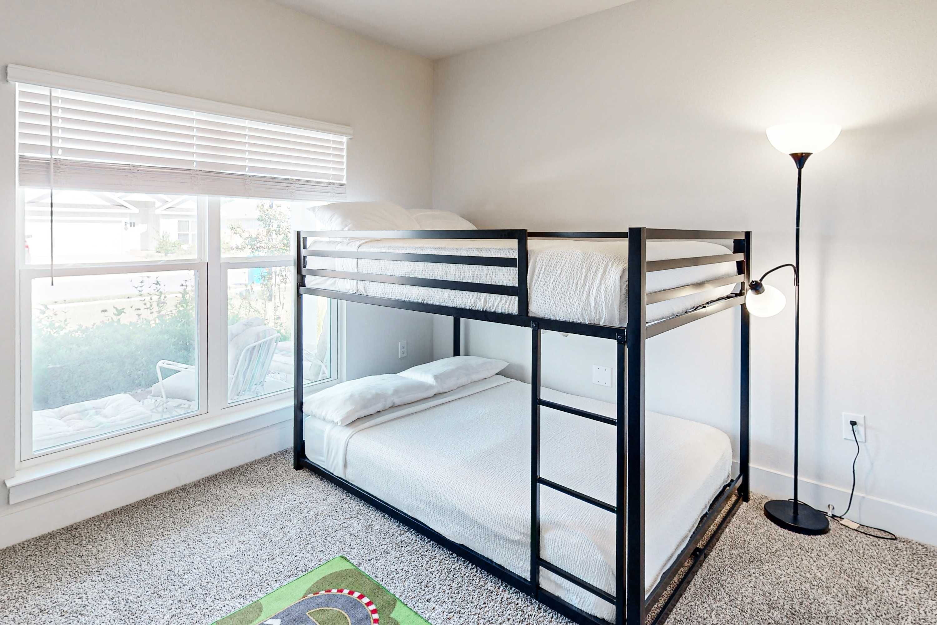 Una cama litera con colchón y almohadas blancos, una ventana con persianas, una lámpara de pie y una alfombra con diseño circular.