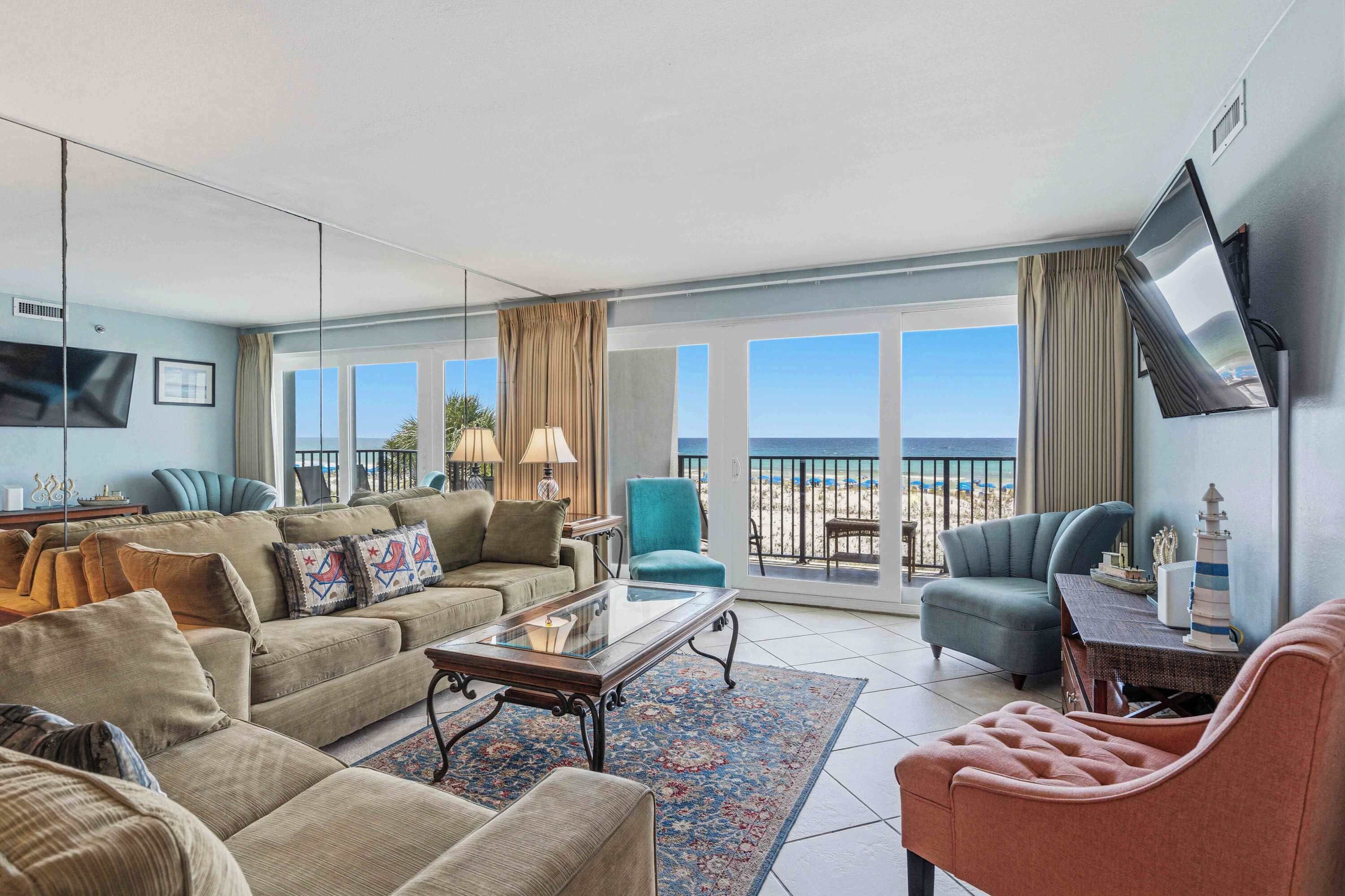 Un salon spacieux avec un grand canapé, une table basse et un téléviseur fixé au mur. Il y a un balcon avec vue sur la plage.