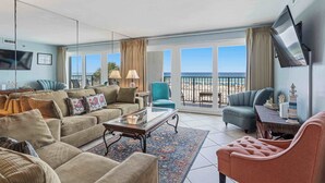 Ein geräumiges Wohnzimmer mit einer großen Couch, einem Couchtisch und einem an der Wand befestigten Fernseher. Es gibt einen Balkon mit Blick auf den Strand.