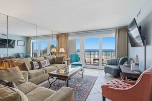 Ein geräumiges Wohnzimmer mit einer großen Couch, einem Couchtisch und einem an der Wand befestigten Fernseher. Es gibt einen Balkon mit Blick auf den Strand.