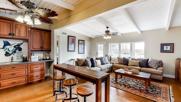 A spacious living area with a high ceiling, wooden ceiling beams, a large window, a sofa set, a coffee table, and a dining area with stools. - Dog-friendly beach home with expansive deck, amazing views, & outdoor shower (Pensacola Beach)
