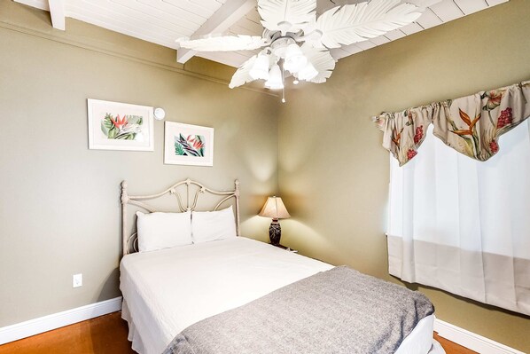 Een slaapkamer met een bed, een plafondventilator, decoratieve muurkunst en een raam met gordijnen.