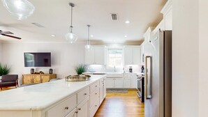 Una cocina moderna con armarios blancos, una isla grande, iluminación colgante y un televisor de pantalla plana montado en la pared.