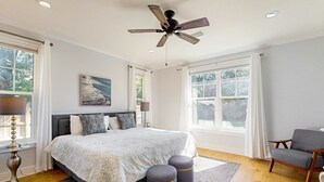 Un dormitorio con una cama grande, un ventilador de techo, una ventana con cortinas, una silla y una lámpara.