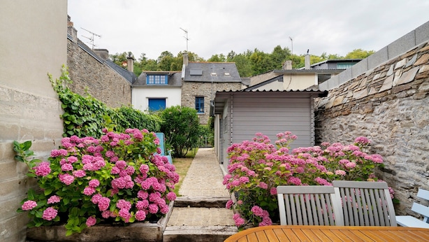 Un estrecho callejón con paredes de piedra, una mesa de madera y flores rosadas.