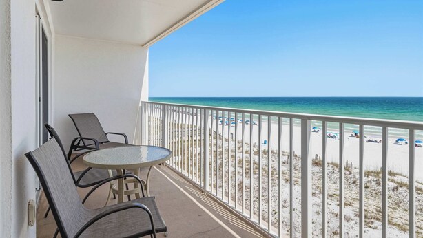 A balcony with a table and chairs, overlooking a beach with umbrellas and a white fence. - Dreamy island escape with beach access, pool & complimentary seasonal beach service (Fort Walton Beach)